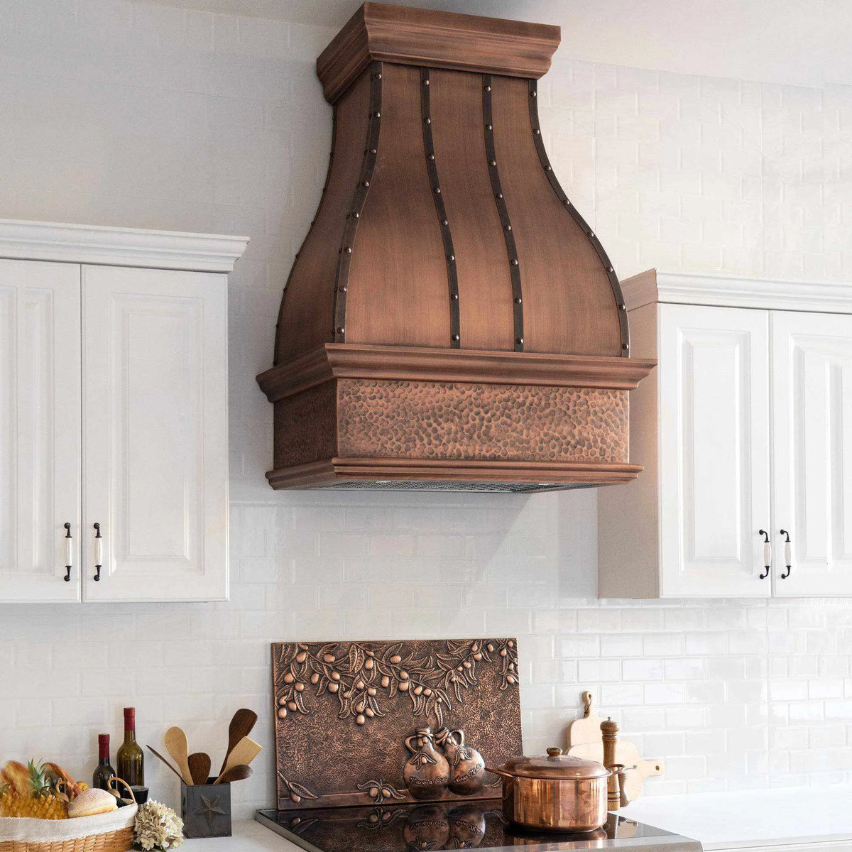 Antique copper range hood with a hammered bottom apron, installed above a kitchen counter, featuring rivets and a bell design.