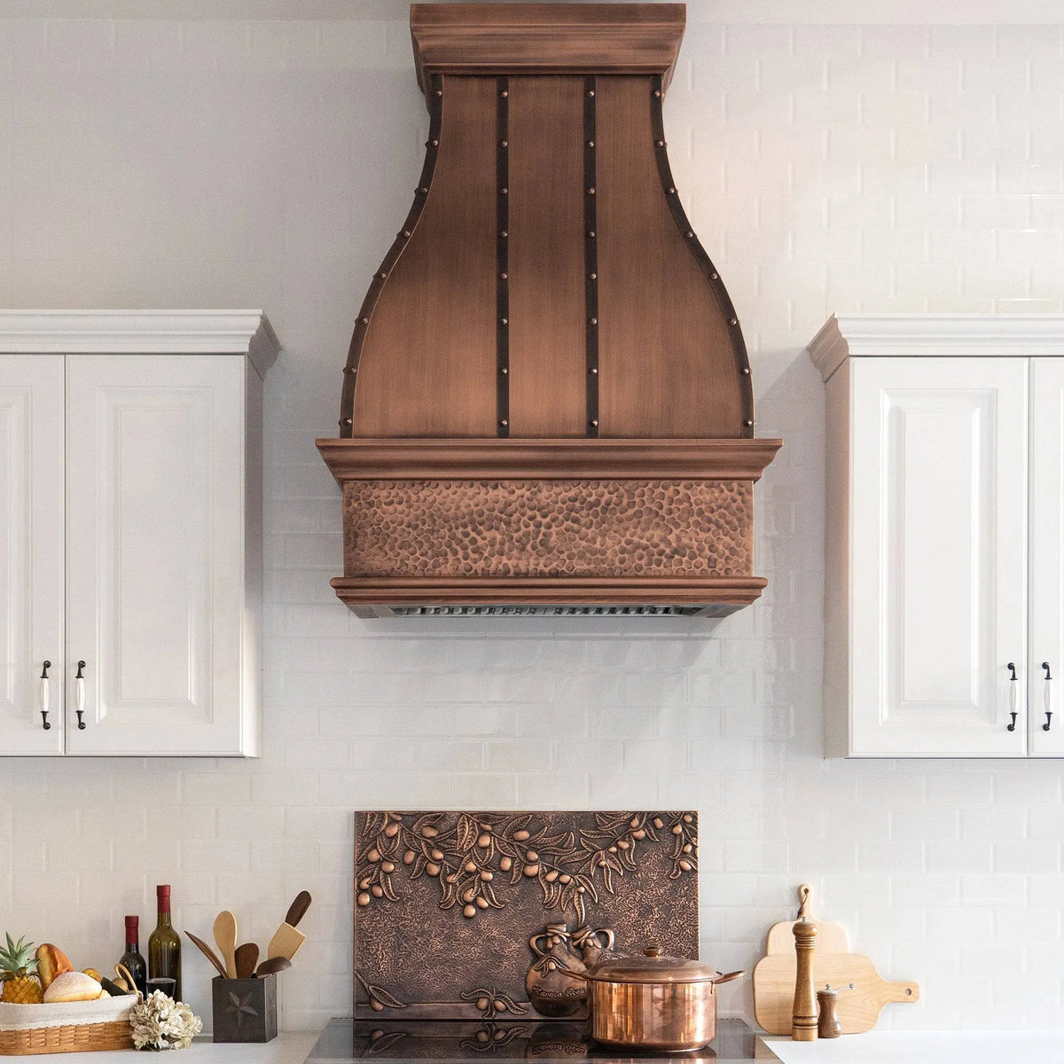 Antique copper range hood with a hammered bottom apron, installed above a kitchen counter, featuring rivets and a bell design.