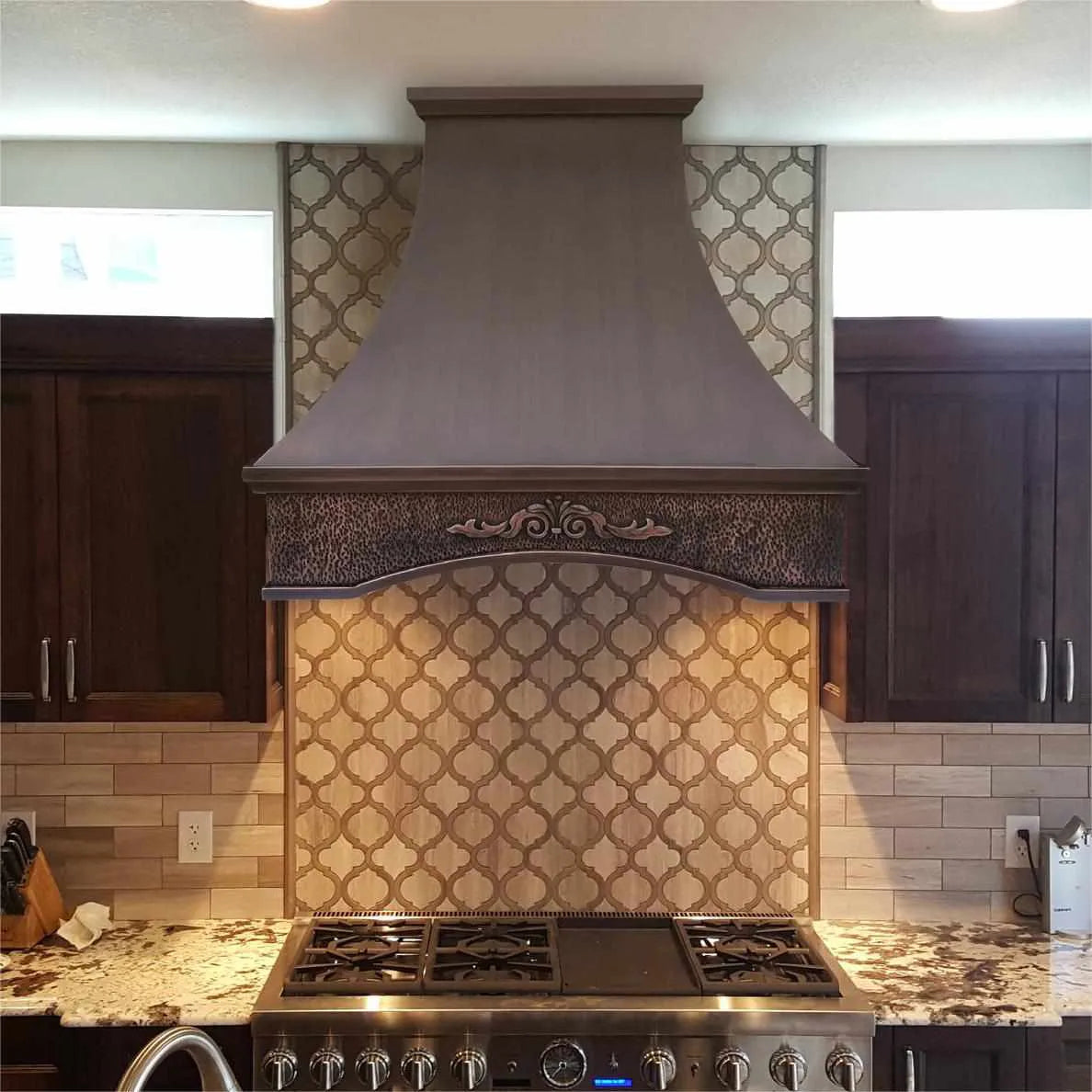 An elegant Fobest copper range hood with a center apron installed over a kitchen stove, featuring a dark antique copper finish.