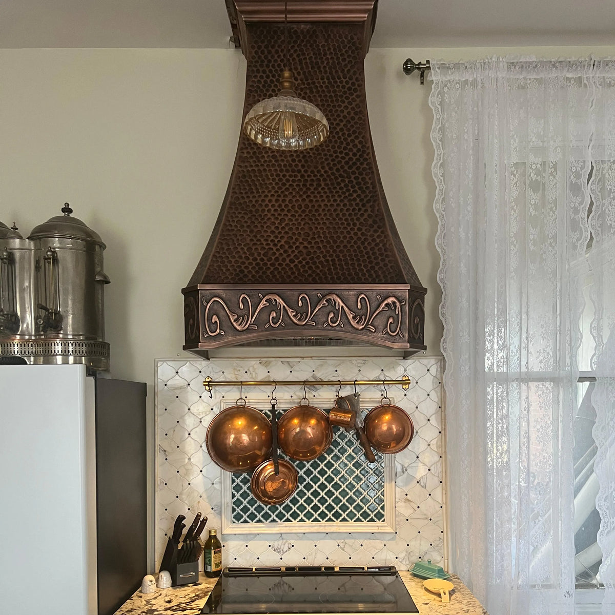 Antique copper range hood with a classic apron design, installed above a kitchen stove with pots hanging below it.
