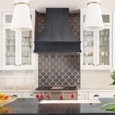 A handcrafted custom copper range hood with a weathered bronze finish, mounted above a kitchen stove against a wall with fishscale tile pattern.
