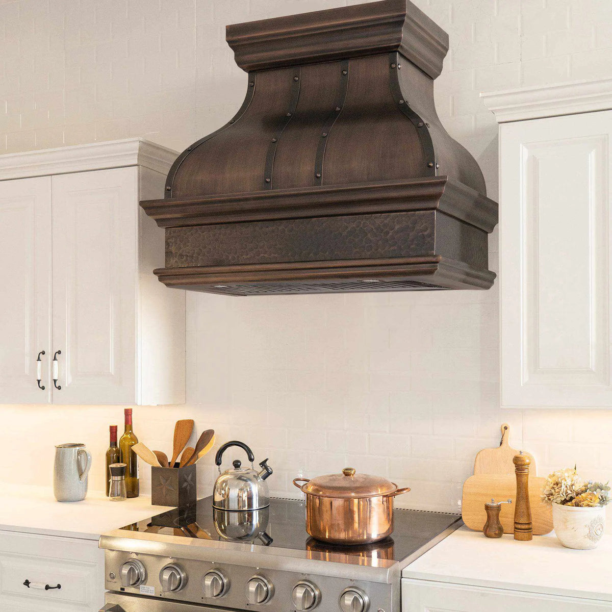 A dark antique copper range hood with a bell shape design, mounted above a kitchen stove against a white tile wall.