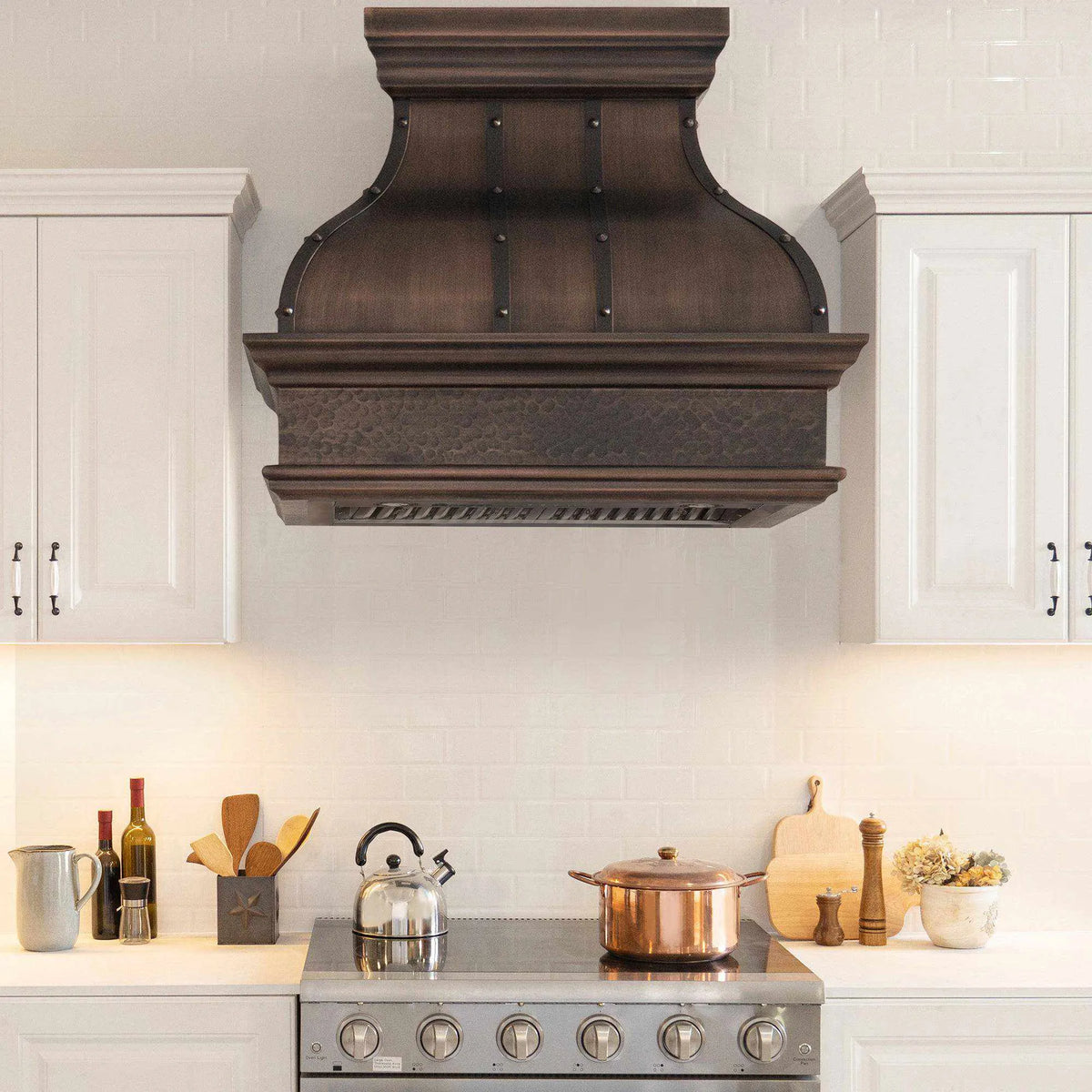 A dark antique copper range hood with a bell shape design, mounted above a kitchen stove against a white tile wall.