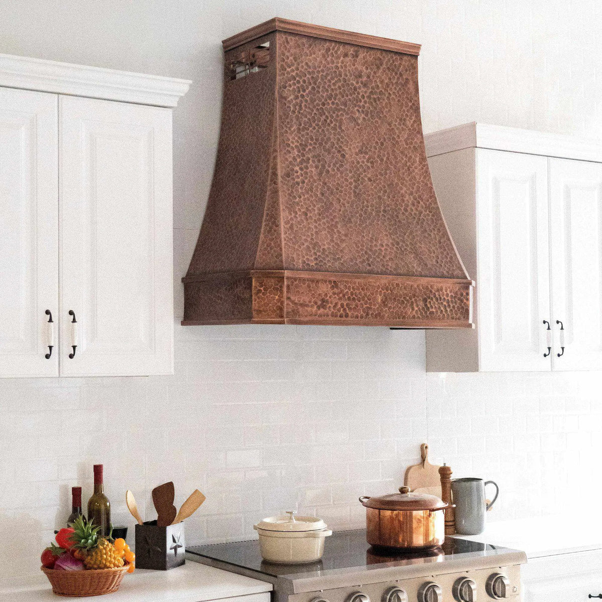 Antique copper stove hood installed above a white cabinet kitchen with light hammered texture design