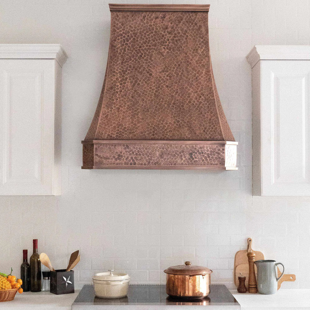 Antique copper stove hood installed above a white cabinet kitchen with light hammered texture design