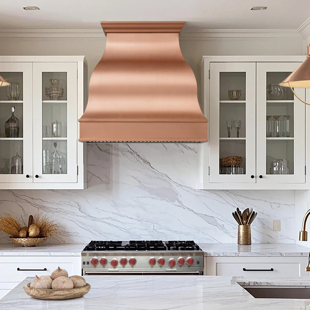A copper range hood with a wavy design, installed above a kitchen stove in a kitchen with white marble countertops and wooden cabinets.