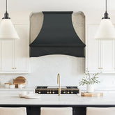 An oil rubbed bronze copper range hood with an arched bottom installed above a kitchen stove, surrounded by white cabinetry and a marble countertop.