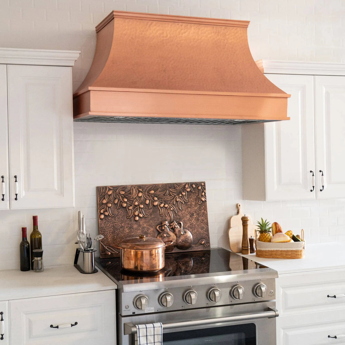 A natural copper range hood with a curved design and light hammered texture, placed above a kitchen stove, with a copper backsplash and kitchen decor in the background.