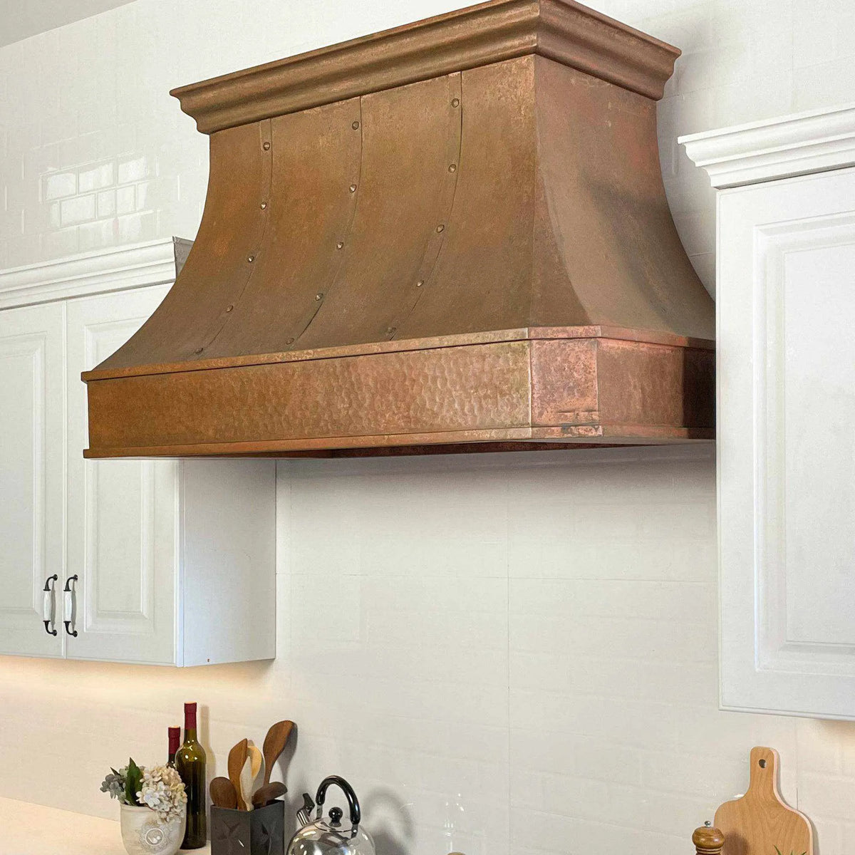 A handmade vintage copper range hood installed above a kitchen stovetop, featuring a curved design with rivets and straps.