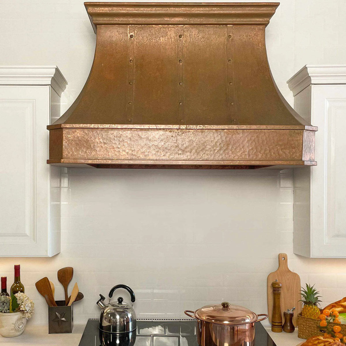 A handmade vintage copper range hood installed above a kitchen stovetop, featuring a curved design with rivets and straps.