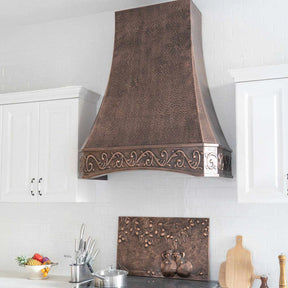 Antique copper range hood with intricate design, installed above a kitchen stove, with vintage apron pattern.
