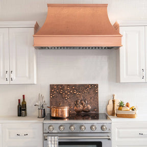 A natural copper range hood with a curved design, placed above a kitchen stove, with a copper backsplash and kitchen decor in the background.