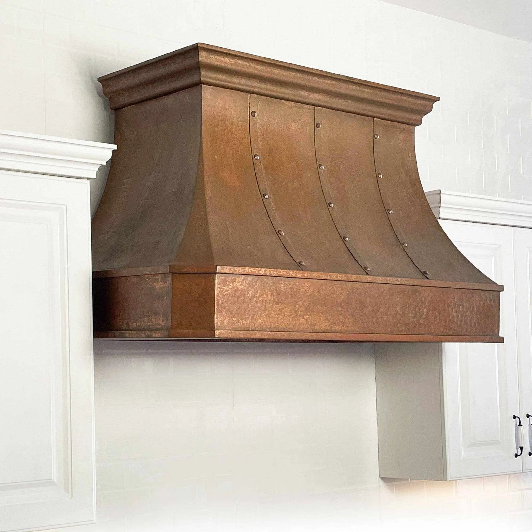 A handmade vintage copper range hood installed above a kitchen stovetop, featuring a curved design with rivets and straps and light hammered texture.