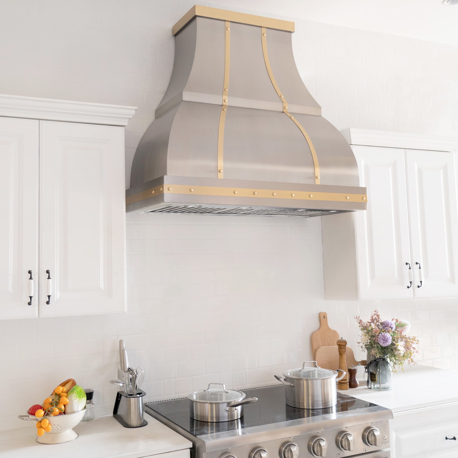 Fobest custom handmade brushed stainless steel range hood with brass straps and rivets installed in the white kitchen